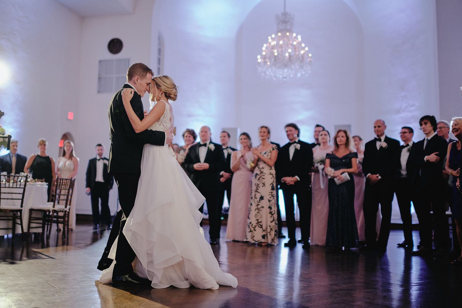 A couple dancing their first dance in an elegant white ballroom while guests watch from the sides.