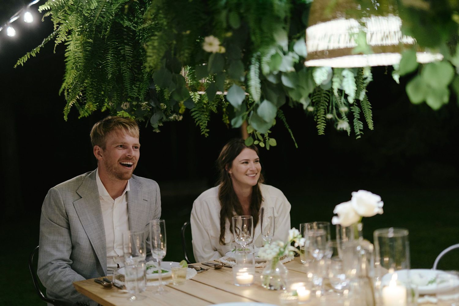 A couple enjoys an elegant outdoor dinner party beneath lush greenery with glowing candles and glassware on the table.