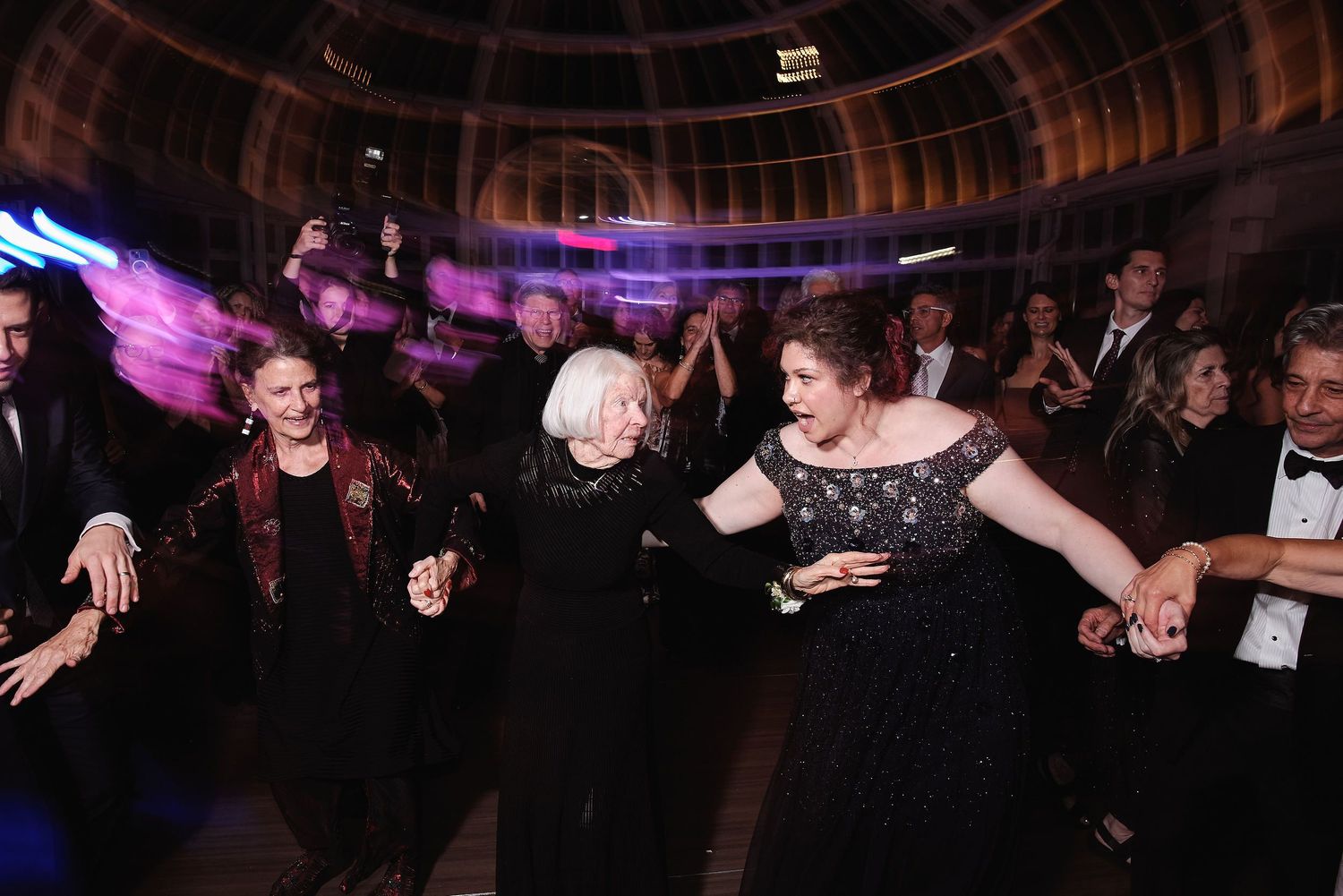 Wedding guests in formal black attire dance and laugh together at an elegant venue with purple lighting.
