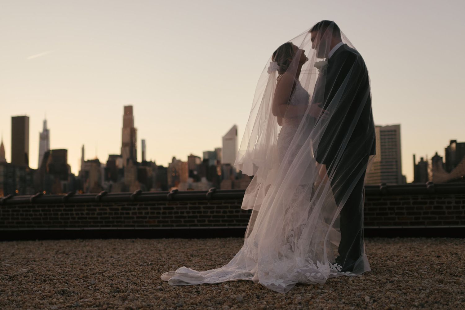 Bride and groom share a romantic kiss under a veil with NYC skyline at sunset.