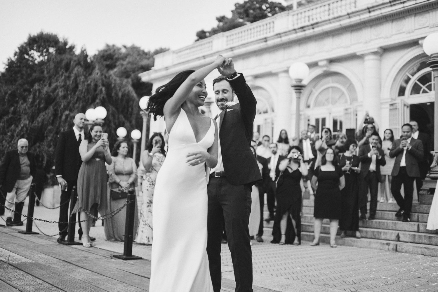 Bride and groom celebrate their wedding exit with raised hands, guests cheering on steps of elegant venue.