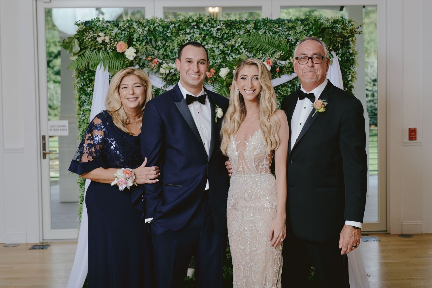A group of four people in formal attire pose together at an elegant event with greenery in the background.