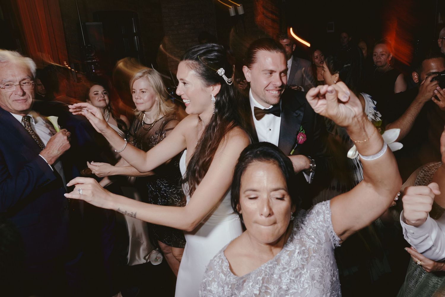 Wedding guests dance and celebrate energetically on a dark dance floor with arms raised during a reception party.