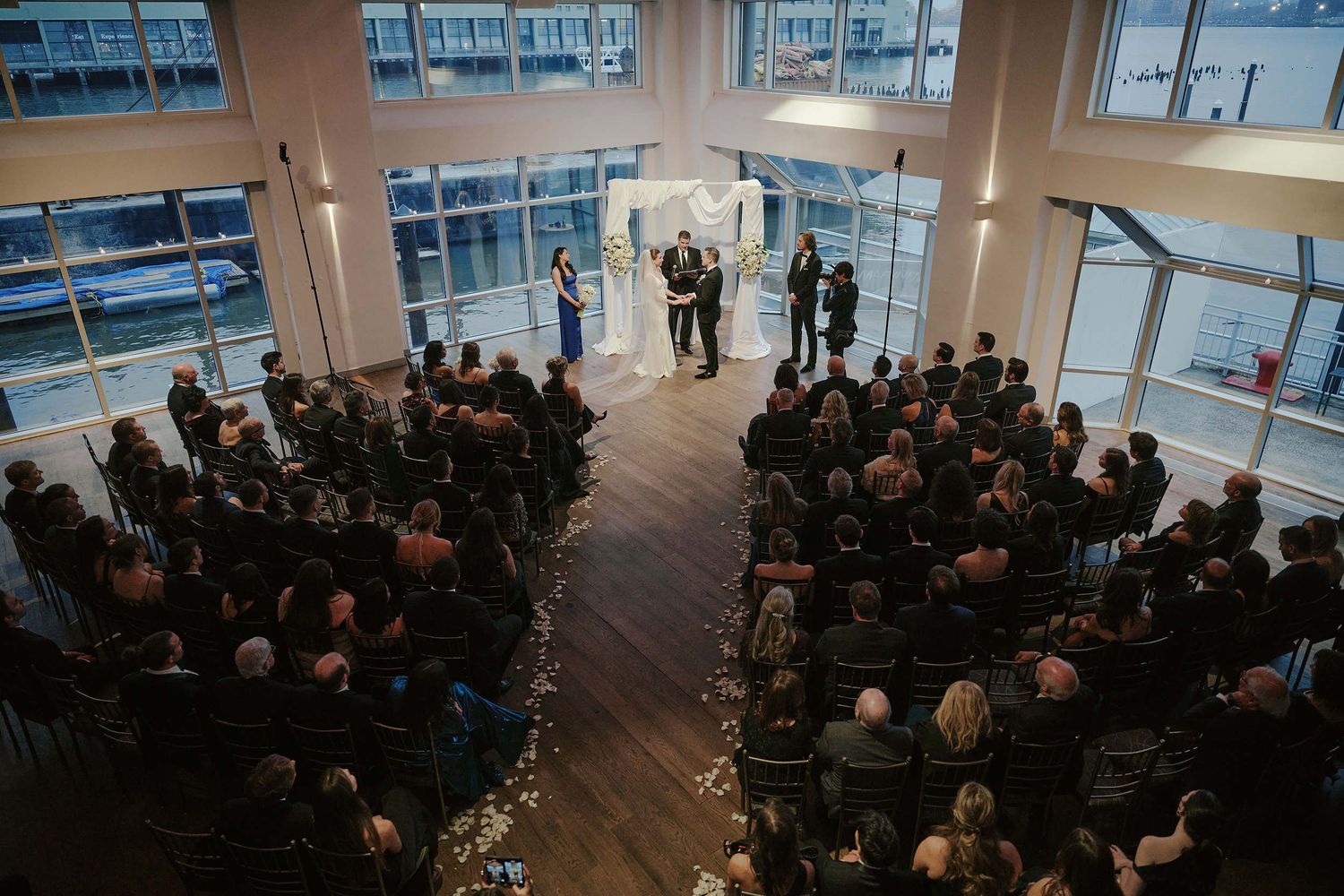 Modern indoor wedding ceremony with guests seated in rows facing glass windows with city views.