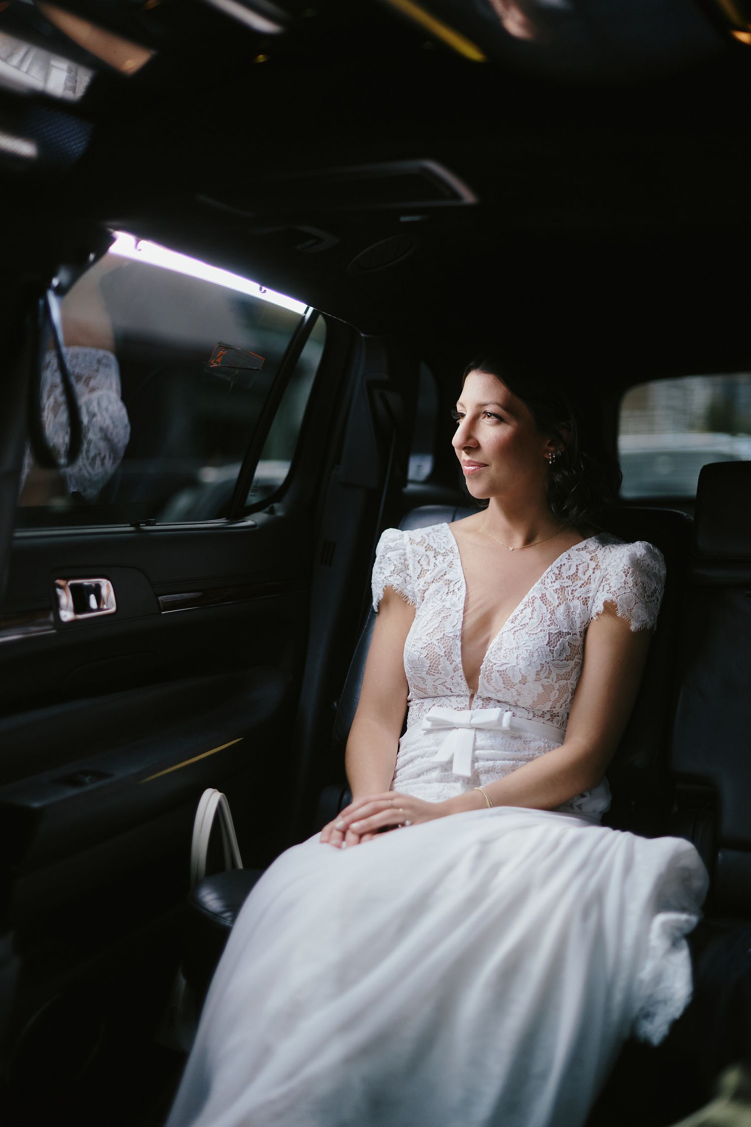 A bride in a delicate lace wedding gown with cap sleeves sits thoughtfully in the backseat of a car.