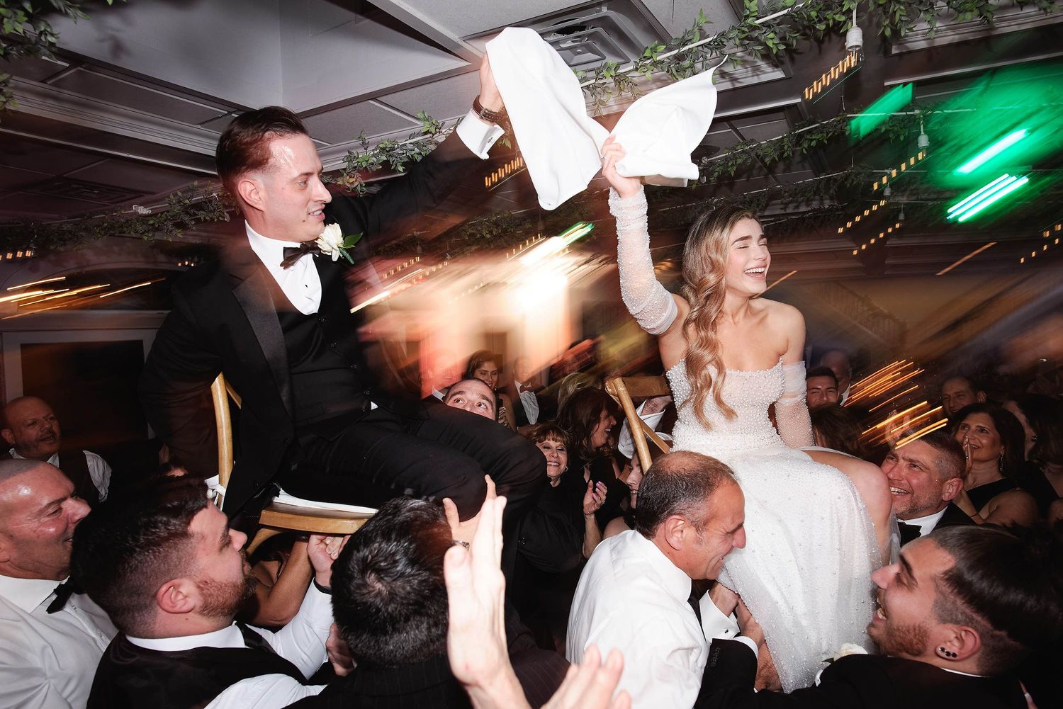Wedding guests lift and celebrate with couple during reception party while throwing napkins in dimly lit venue with green lights.