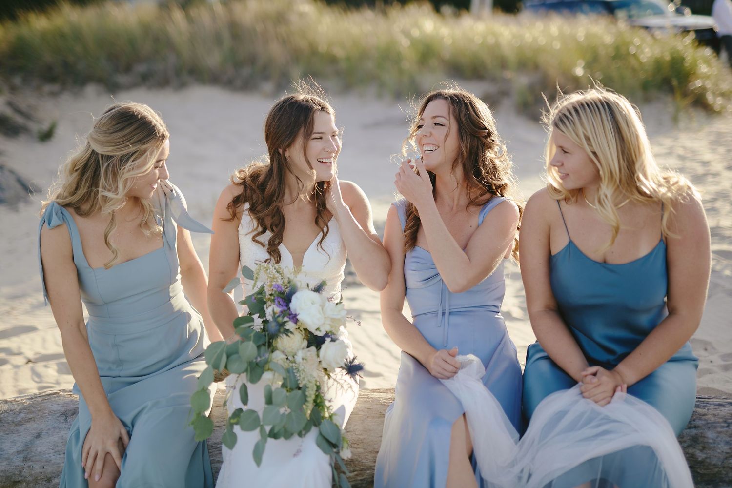 Bridesmaids in light blue dresses sit together outdoors sharing joyful laughter while one holds a floral bouquet.