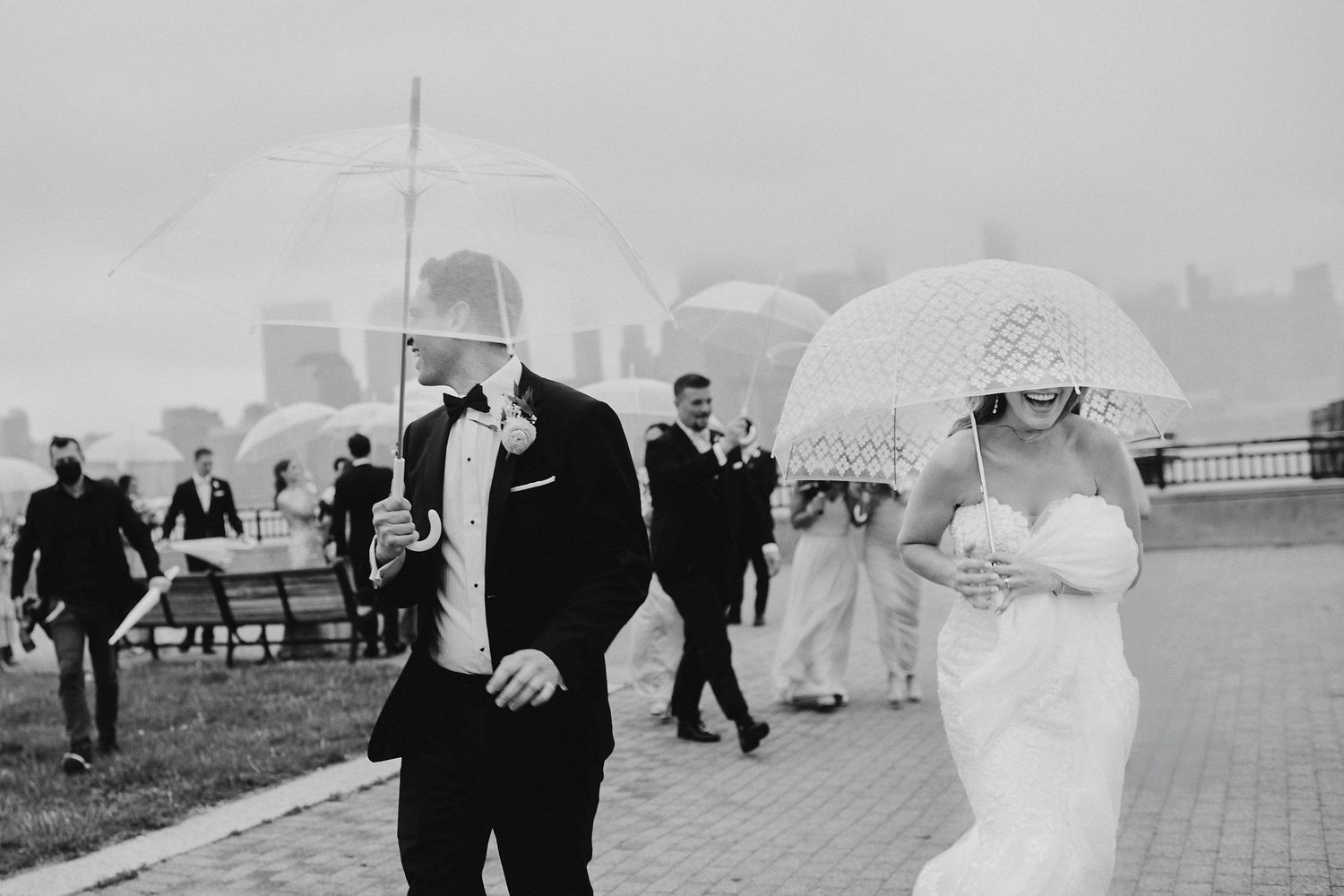 A couple holding clear umbrellas walks together on a brick pathway as wedding guests follow behind them.