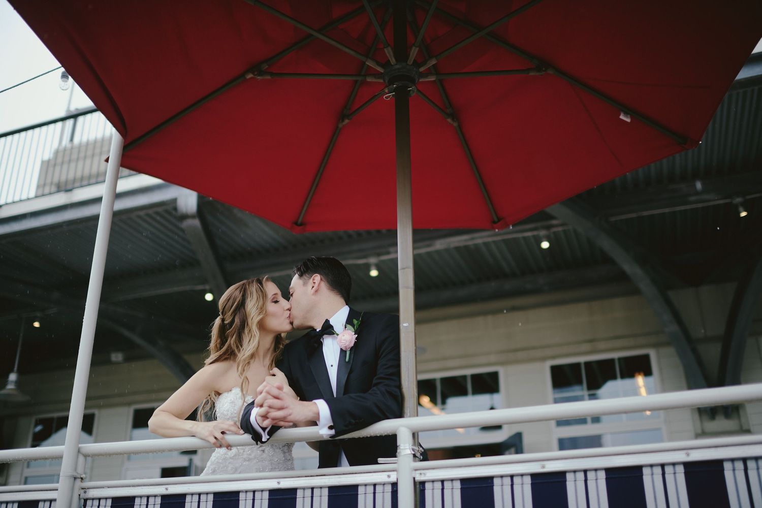 A couple shares a romantic moment under a large red umbrella on a balcony setting.