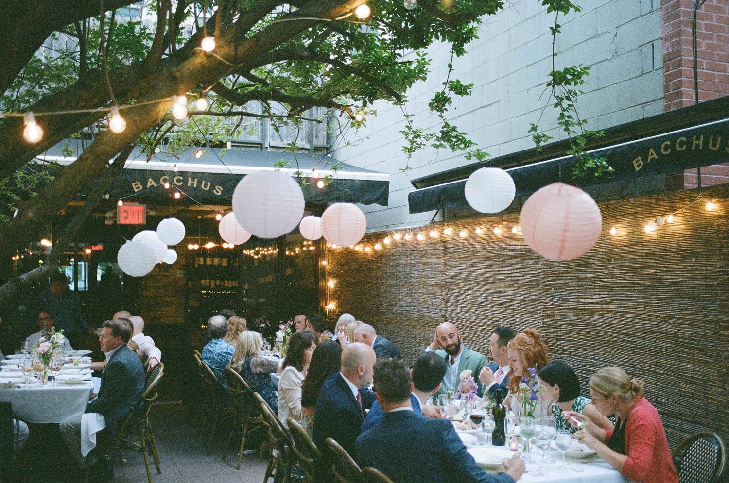Outdoor evening wedding reception with string lights and paper lanterns illuminating guests dining at tables in a garden patio.