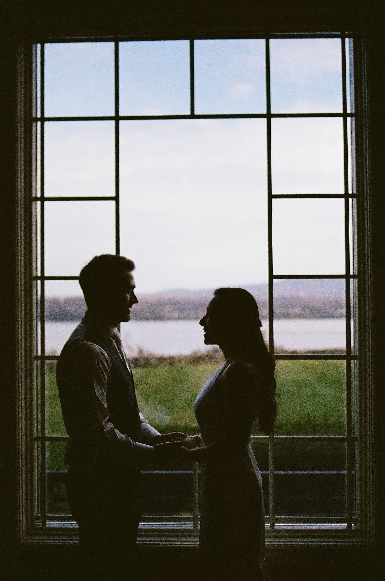 A silhouetted couple stands face to face by a large window with divided panes overlooking a scenic landscape.