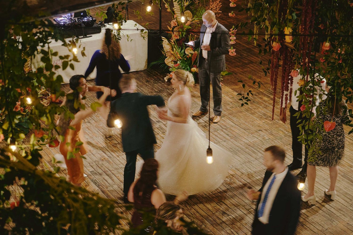 Wedding guests dance together on a wooden deck adorned with twinkling lights and greenery at an evening reception.