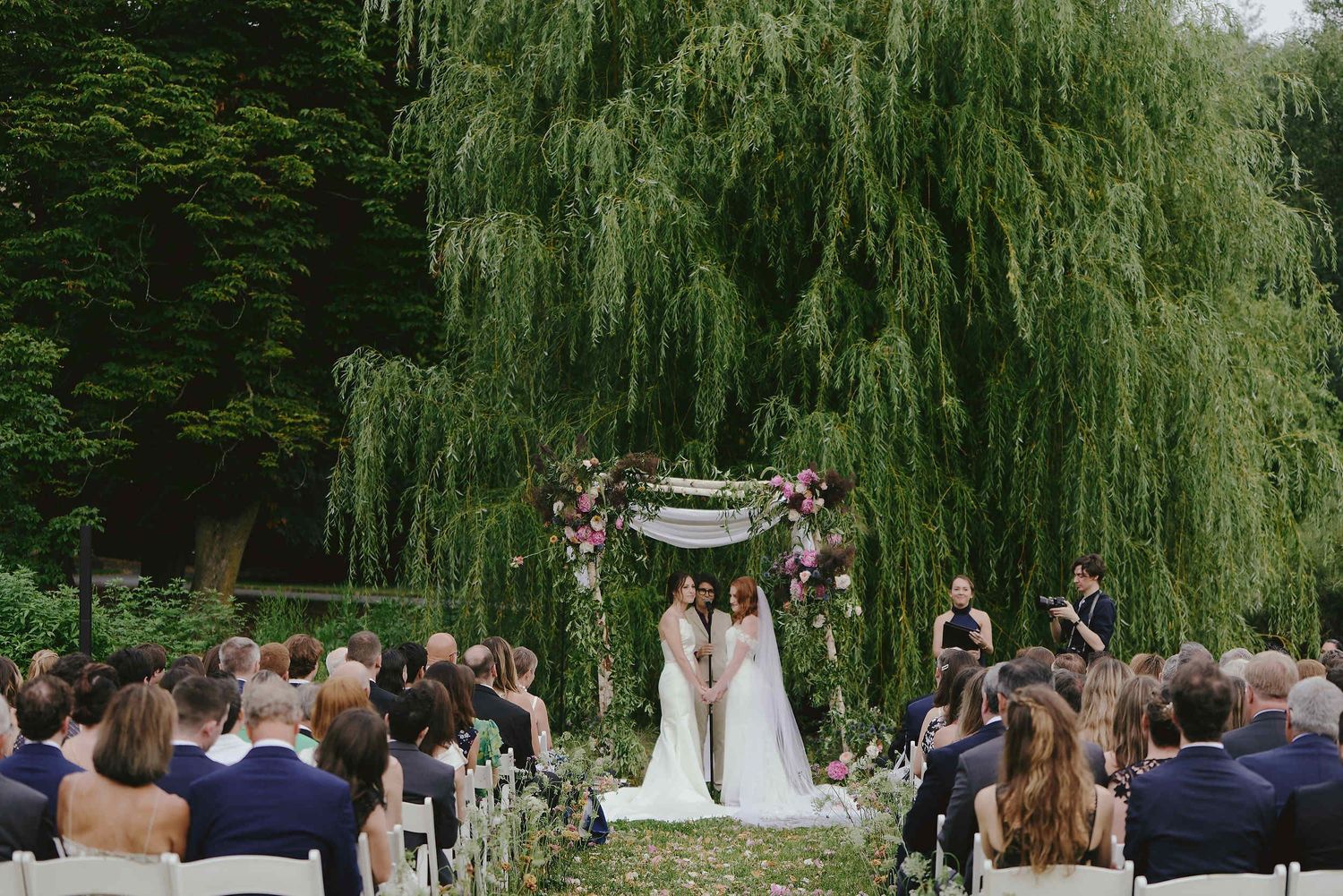 An outdoor wedding ceremony under a floral arch with weeping willows and guests seated on white chairs in a garden setting.
