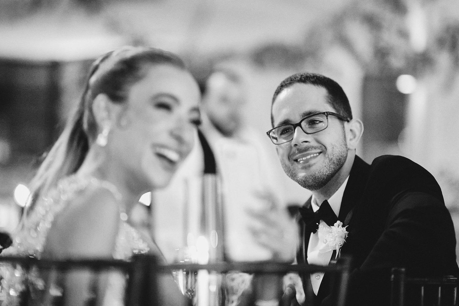 Black and white photo of newlyweds sharing a candid moment during wedding reception speeches.