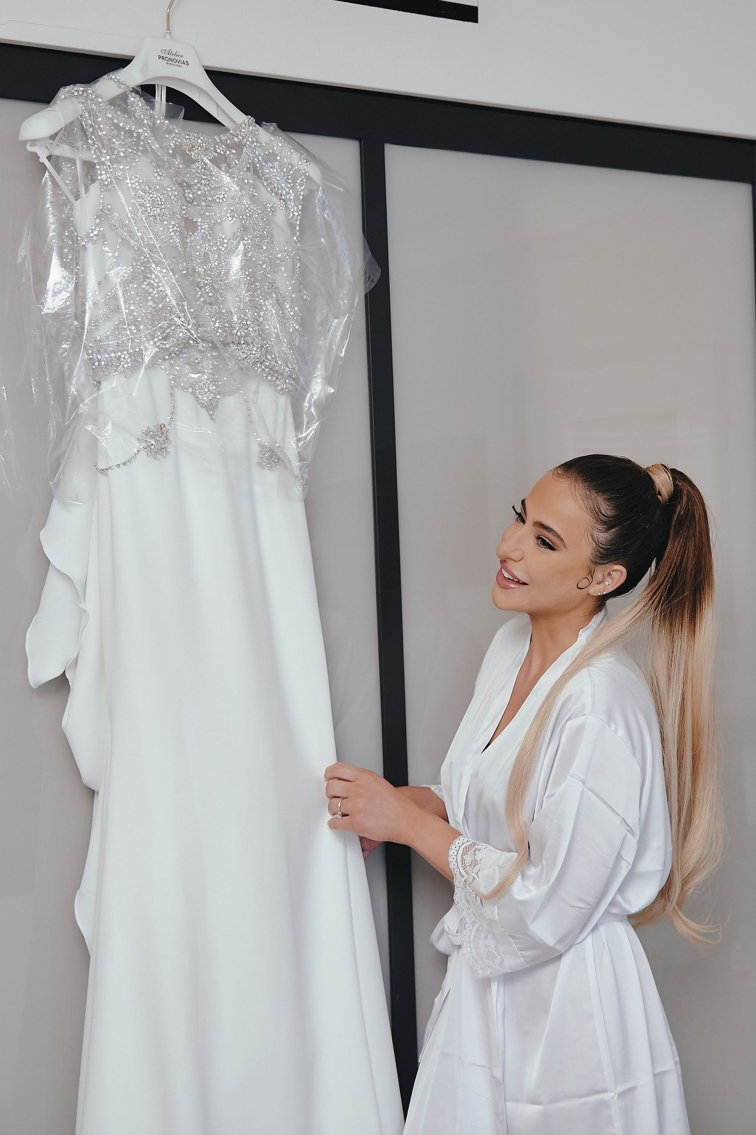 A beautiful white lace wedding dress hangs on display as someone in a white satin robe admires it.