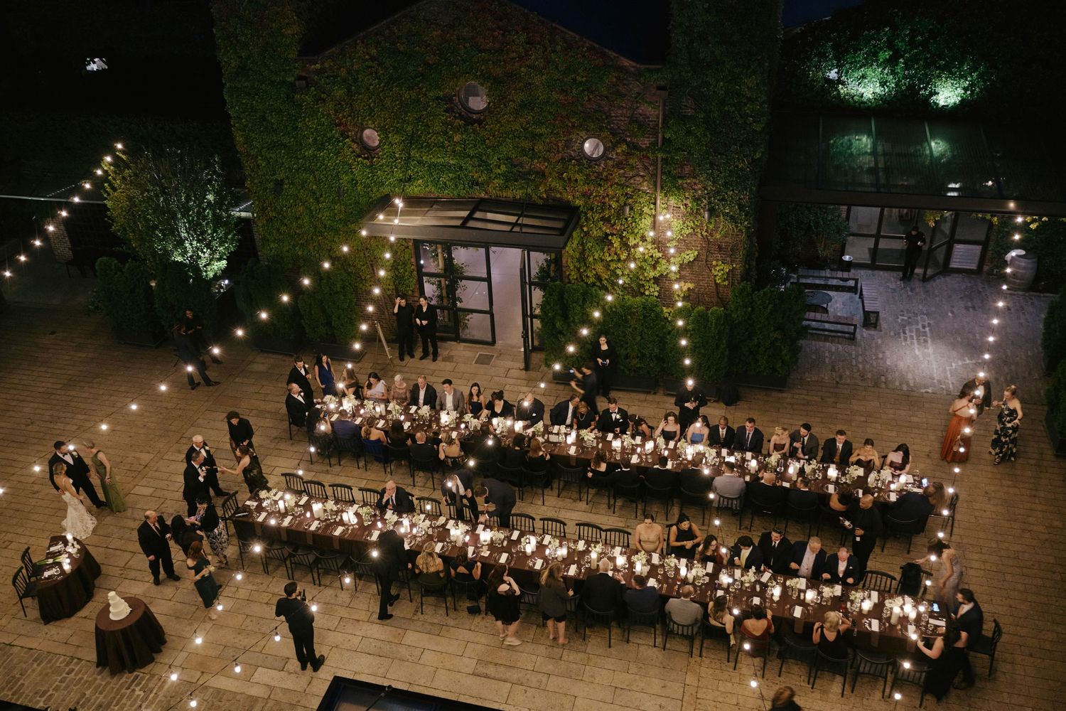 Aerial view of a long outdoor dining table illuminated by string lights during an evening garden party.