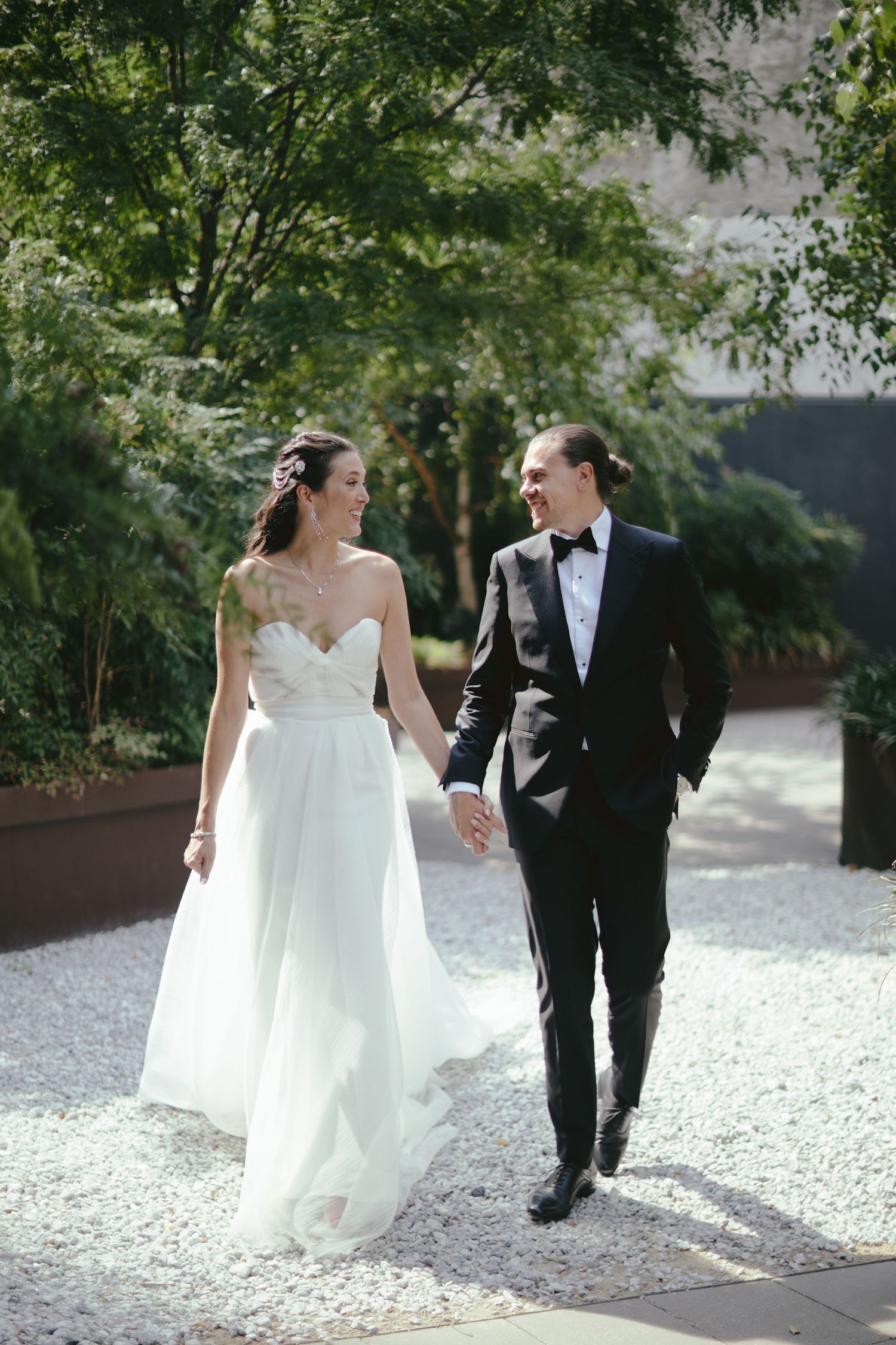 A couple in formal wedding attire walking together on a white gravel path surrounded by lush green trees.