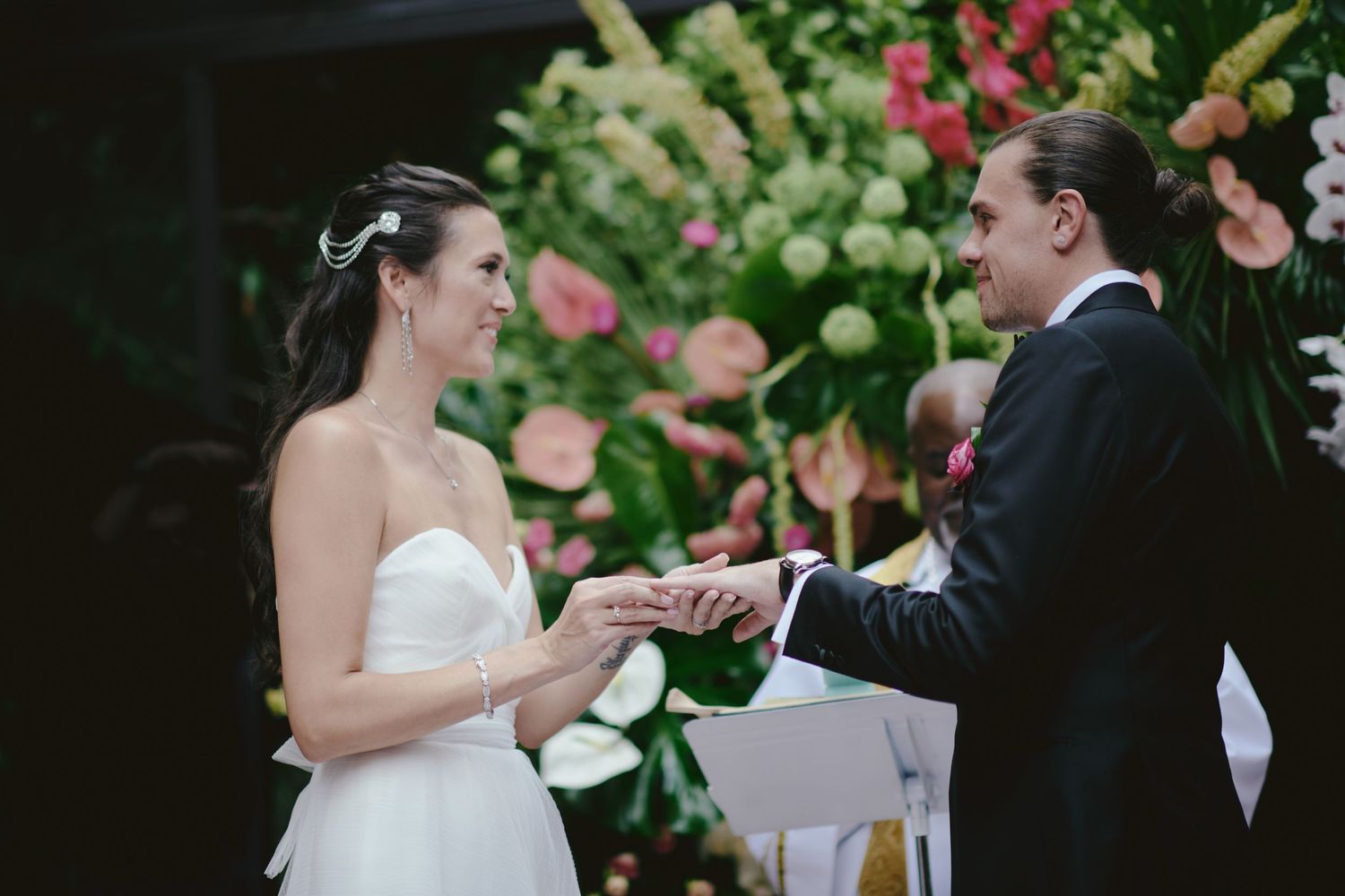 A couple exchanges rings during an outdoor wedding ceremony surrounded by colorful floral arrangements.