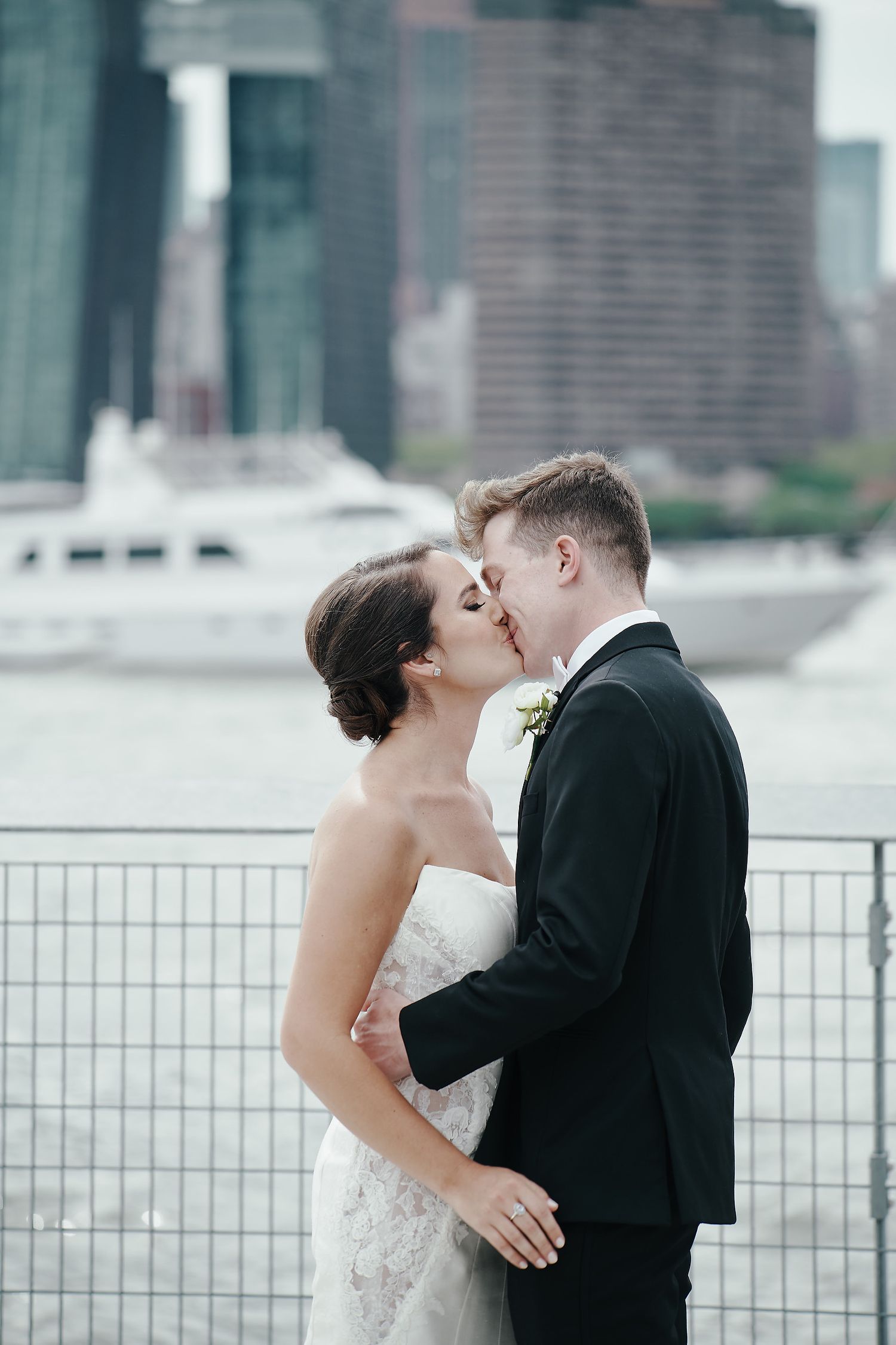A romantic wedding couple sharing a kiss by the waterfront with city buildings in the background.