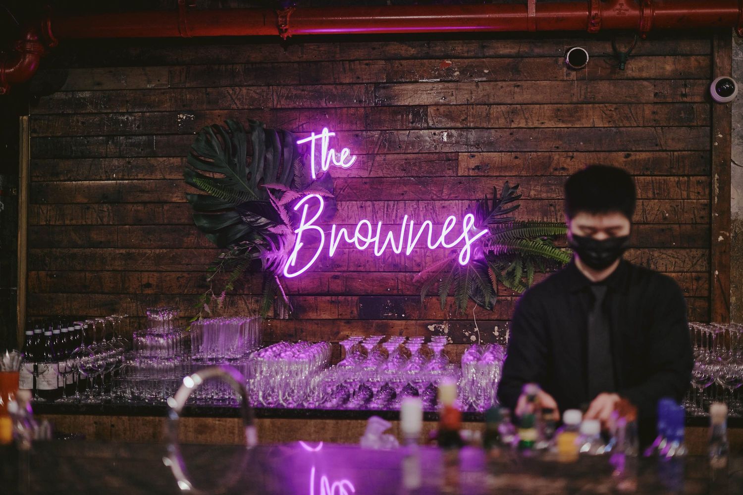 Illuminated pink neon sign reading 'The Brownies' glows against dark brick wall behind bartender at counter.