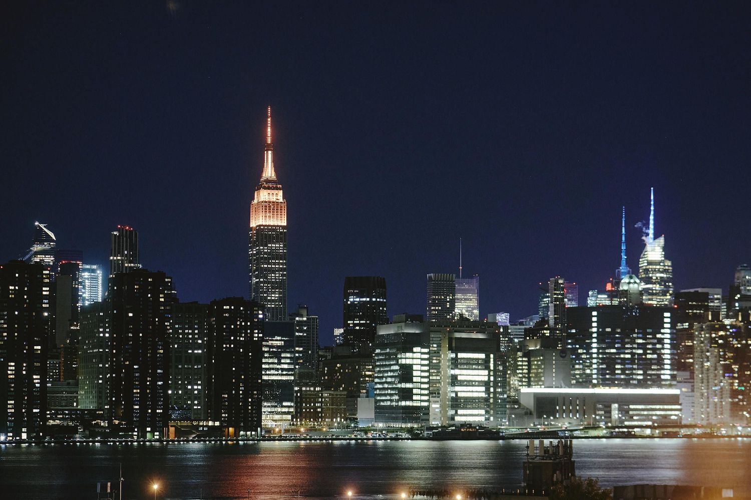 Manhattan skyline illuminated at night with Empire State Building and city lights reflecting on East River.