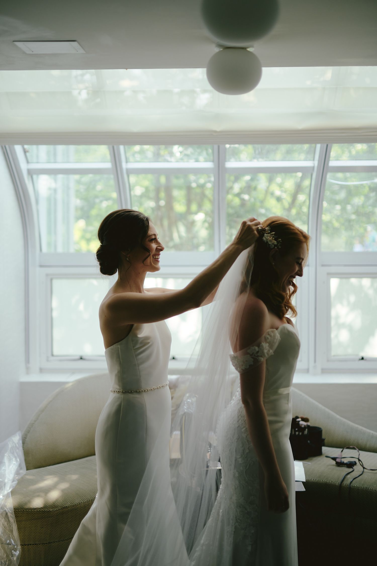 Two people in elegant white dresses prepare for a wedding ceremony in a sunlit room with large windows.