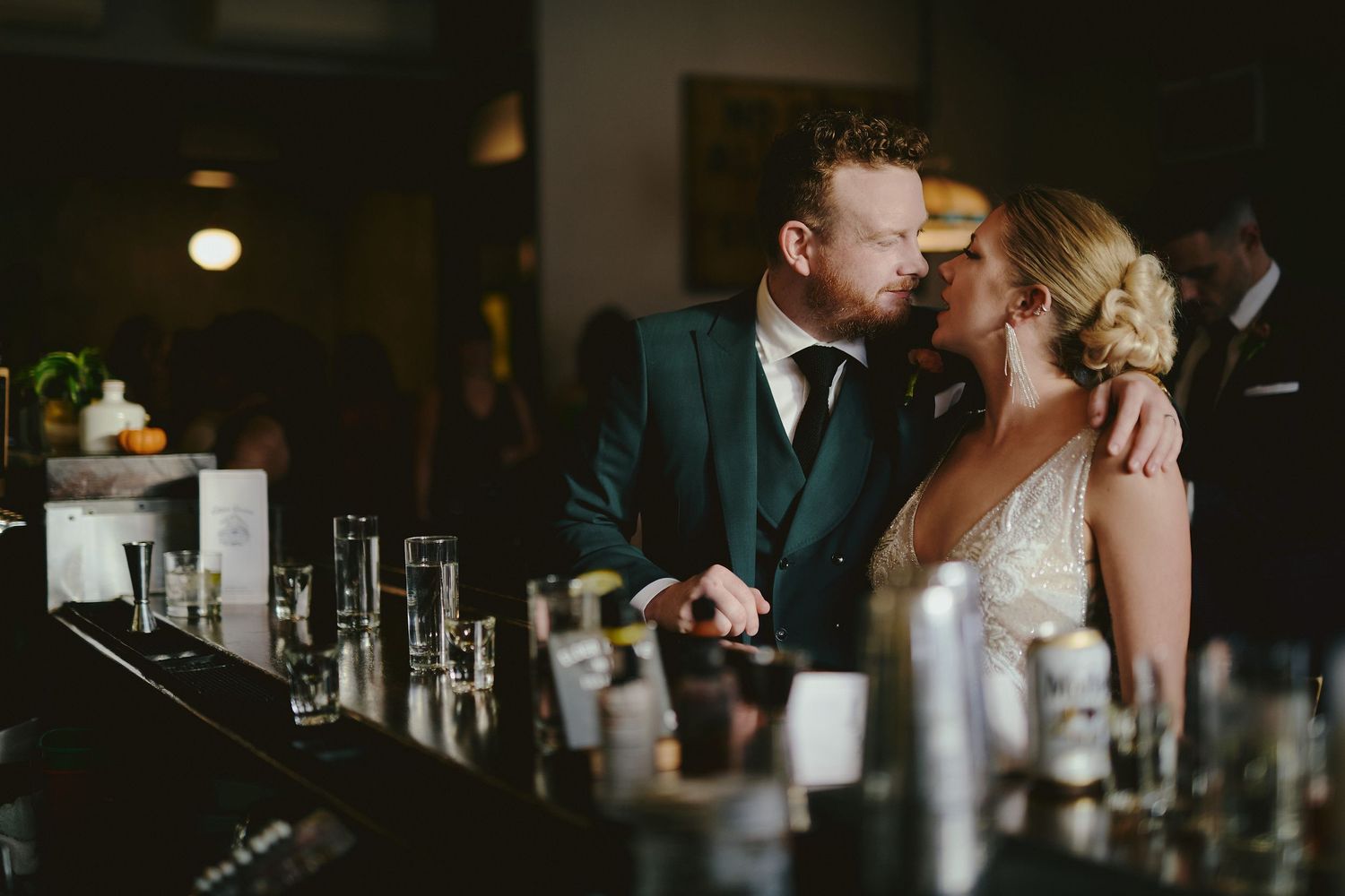 Romantic wedding couple sharing an intimate moment at a dimly lit bar with bottles and glassware in the foreground.