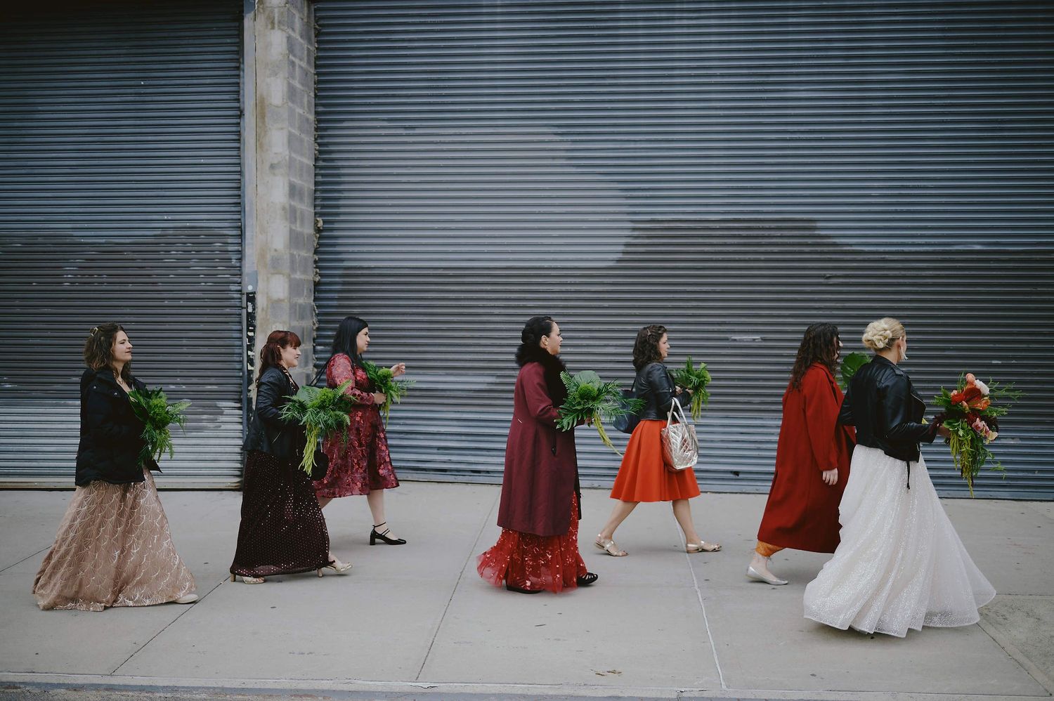 People in long flowing dresses walk in a line carrying greenery and flowers against an industrial backdrop.