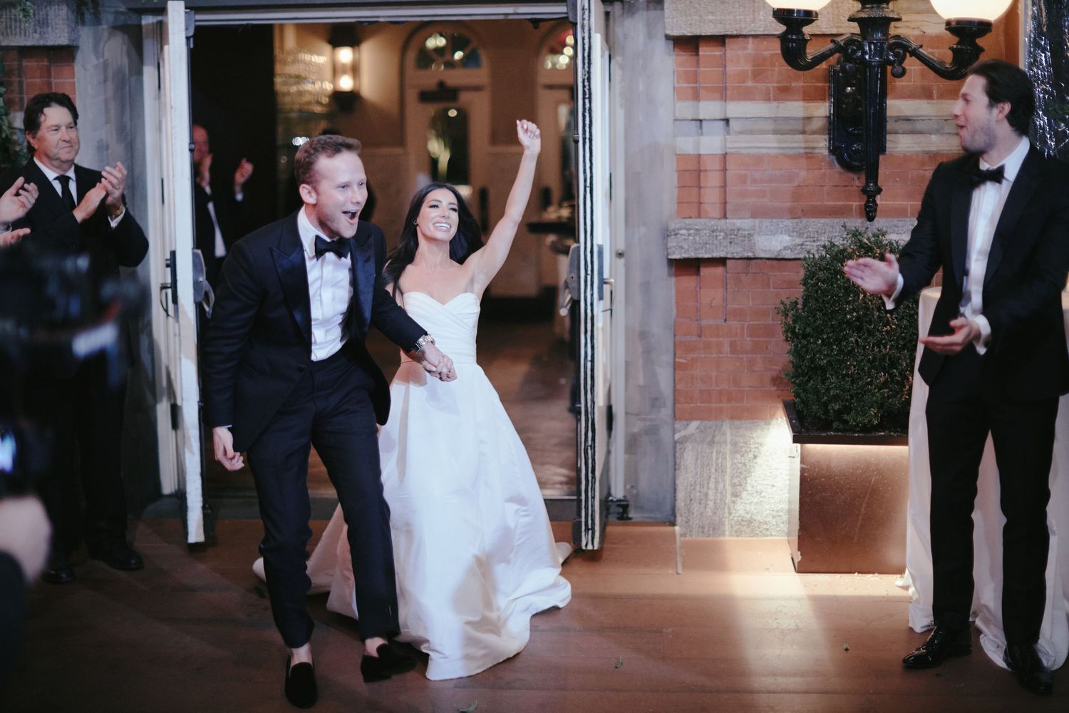 Couple makes joyful entrance at evening wedding reception while guests applaud outside brick venue.