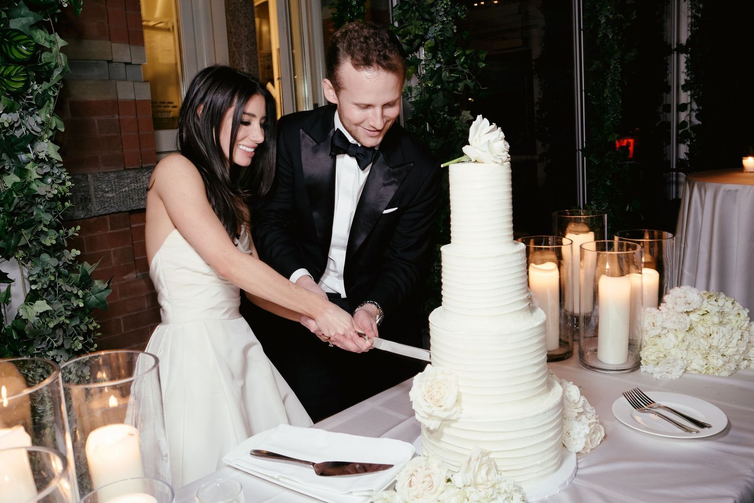 A couple in wedding attire cuts into their elegant white tiered wedding cake surrounded by glowing candles at their reception.