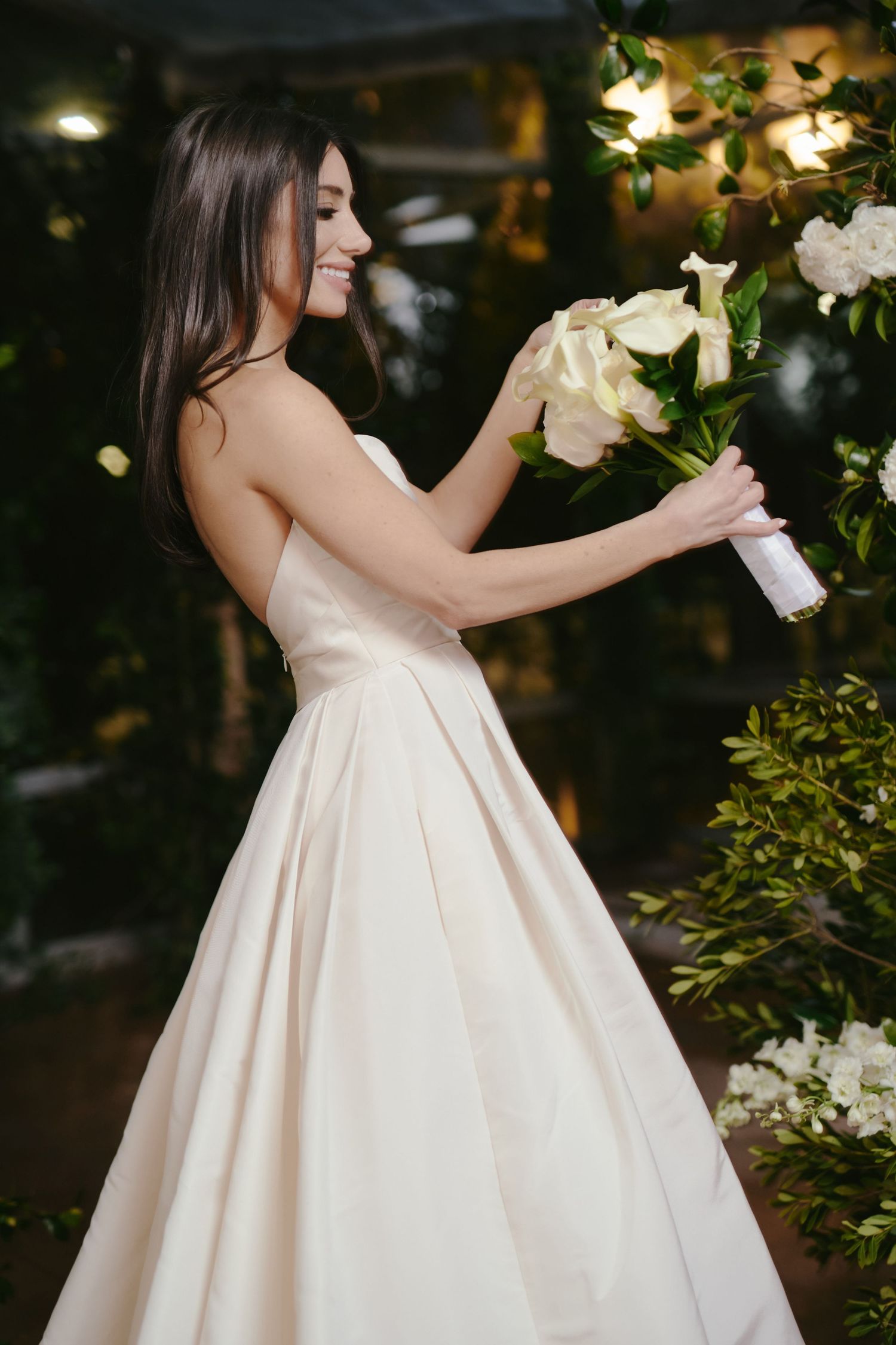 A person in an elegant cream-colored wedding gown arranges white flowers in an evening garden setting.