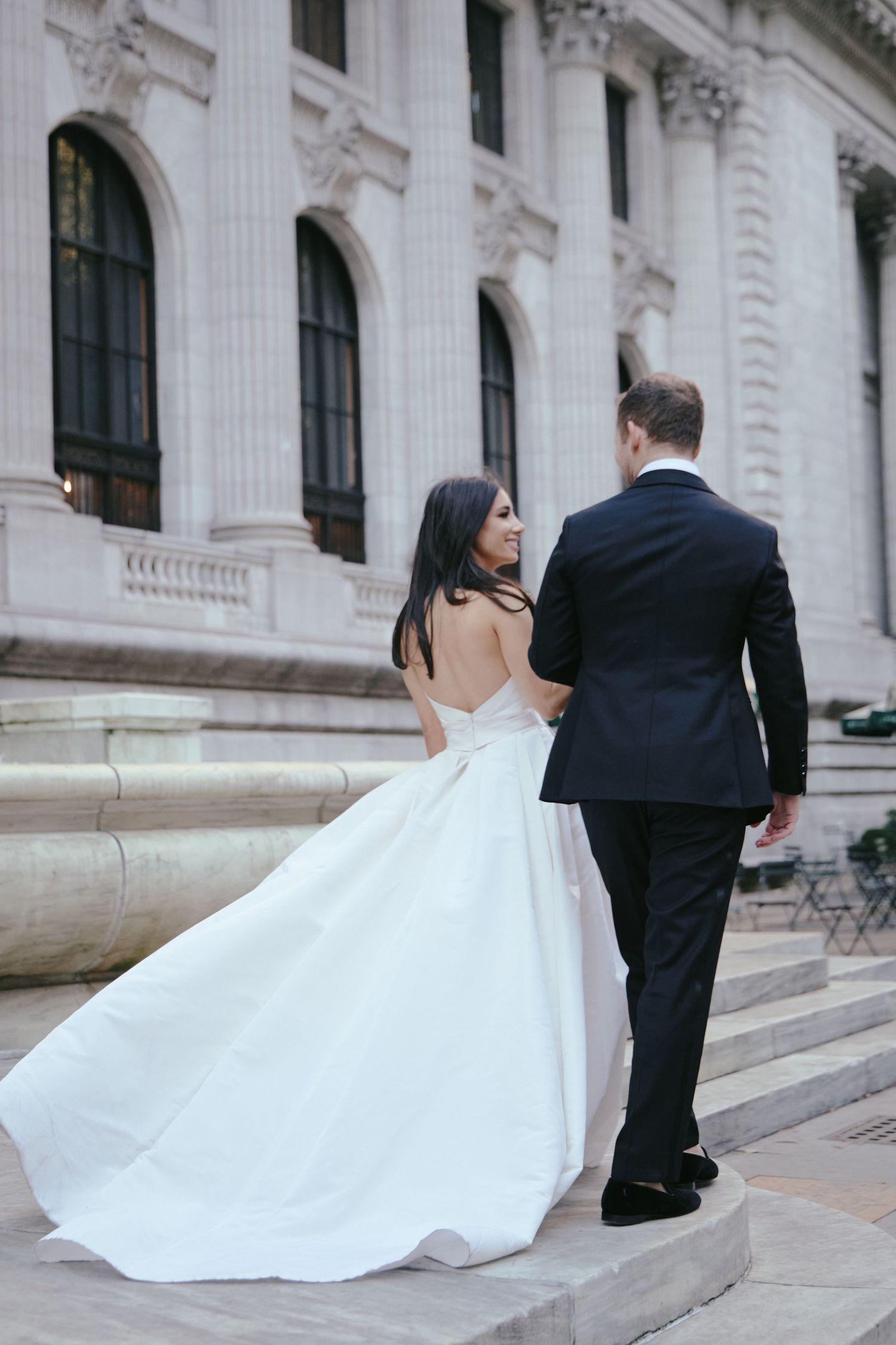 Elegant bride in flowing white ballgown and groom in black tuxedo stand on grand stone steps of classical building.