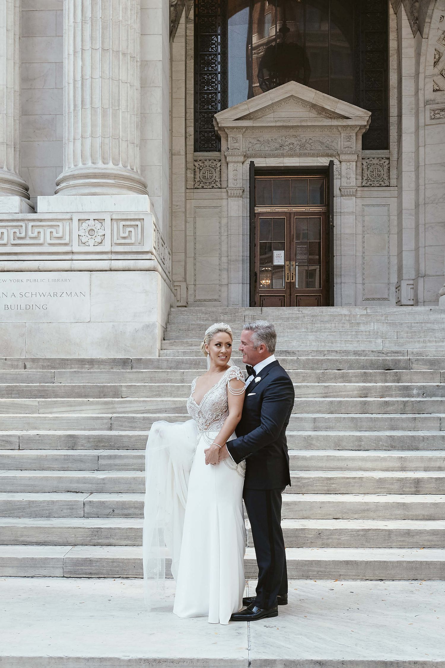 A wedding couple stands on grand stone steps of a classical building with tall columns and ornate architectural details.