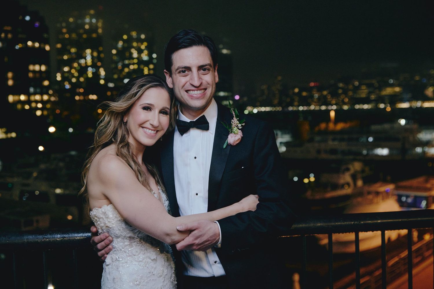 A couple in formal wedding attire embraces on a rooftop terrace with glowing city lights in the background at night.