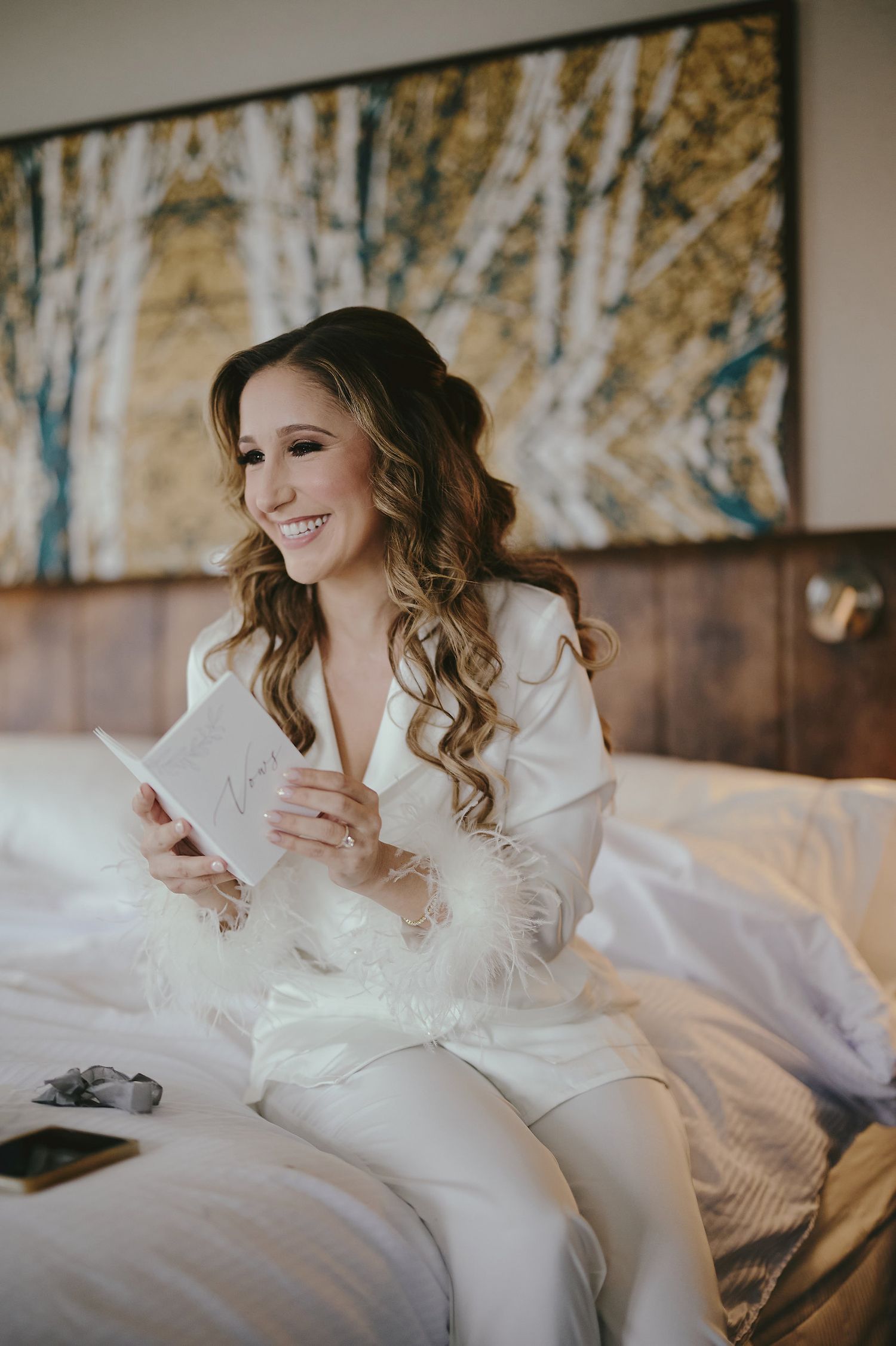 A bride in a white satin robe sits on a bed while reading a letter, with wavy hair and a joyful expression.