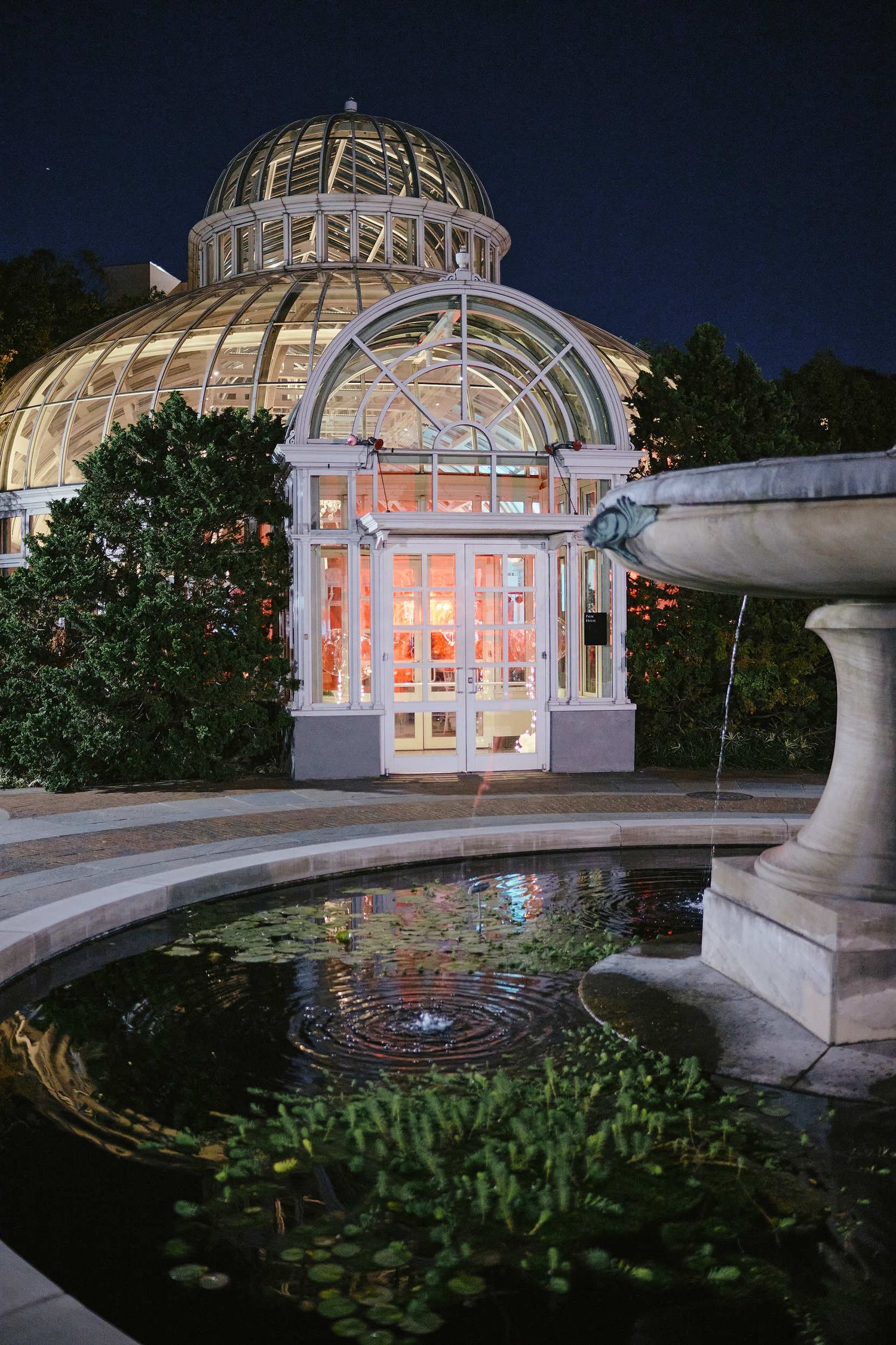 Illuminated glass conservatory with domed roof and reflecting pool at twilight showcases elegant Victorian architecture.