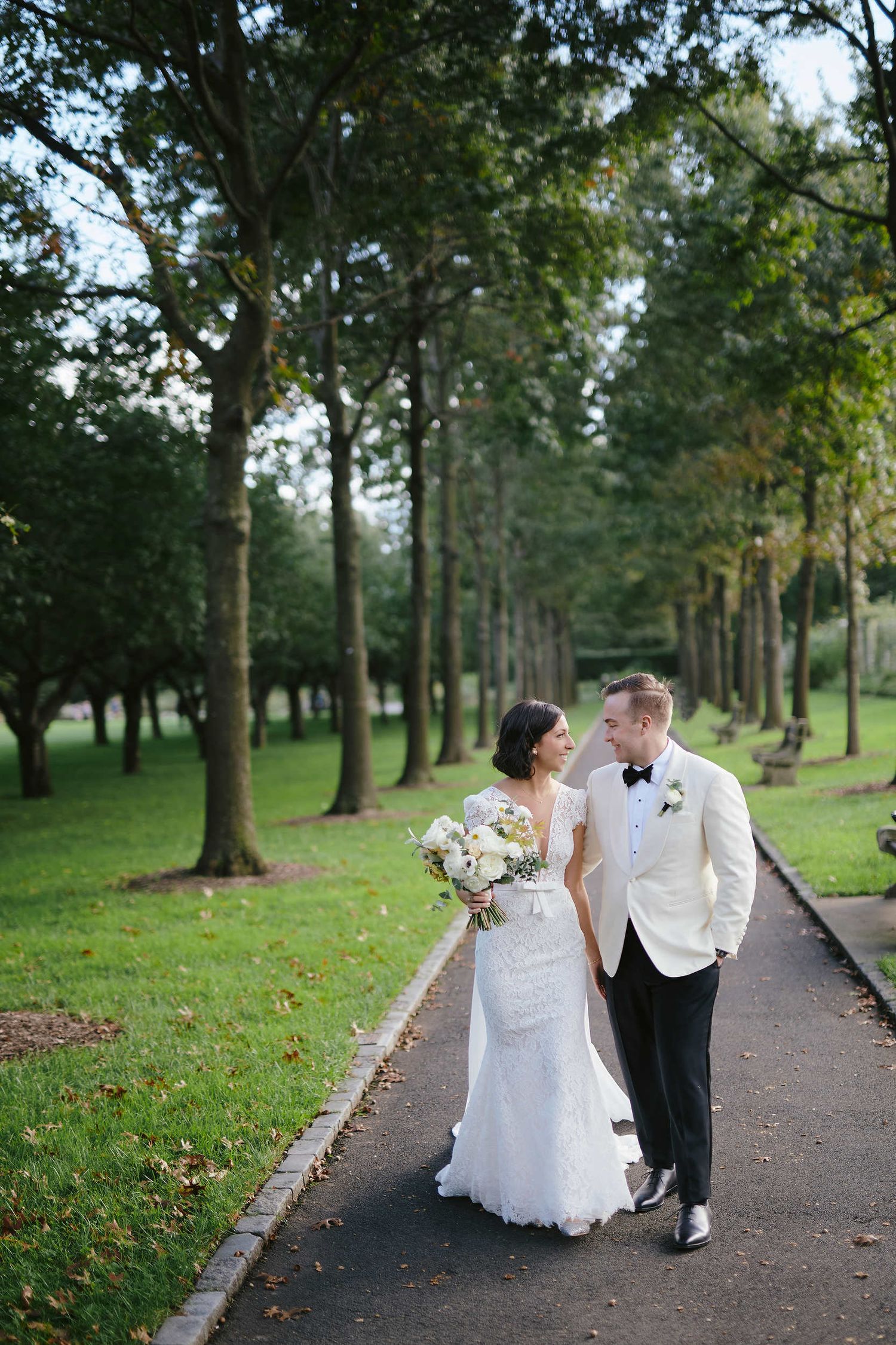 A couple in wedding attire walking together on a tree-lined path in a lush green park setting.