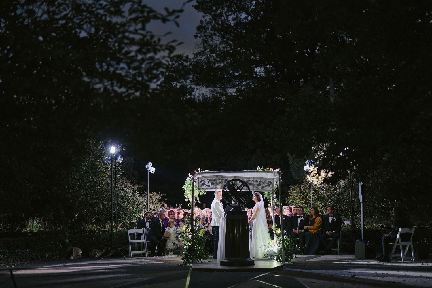 A couple stands under an illuminated wedding arch at night during an outdoor ceremony with guests seated nearby.