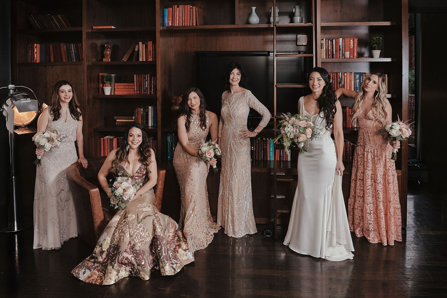 Six bridesmaids in mismatched blush and floral gowns holding bouquets pose elegantly in a wood-paneled library.