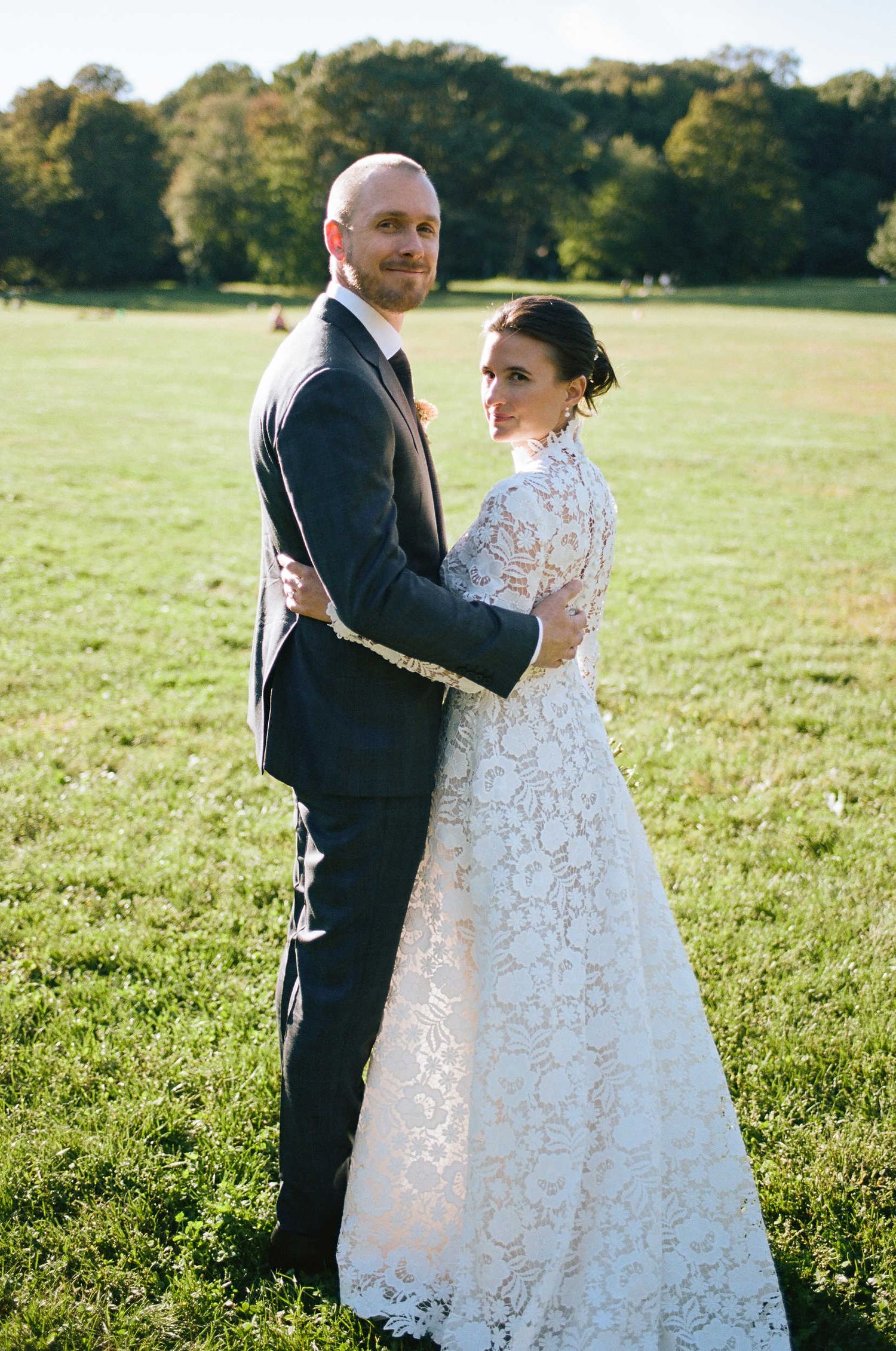A couple poses in formal wedding attire on a sunny lawn with trees in the background.