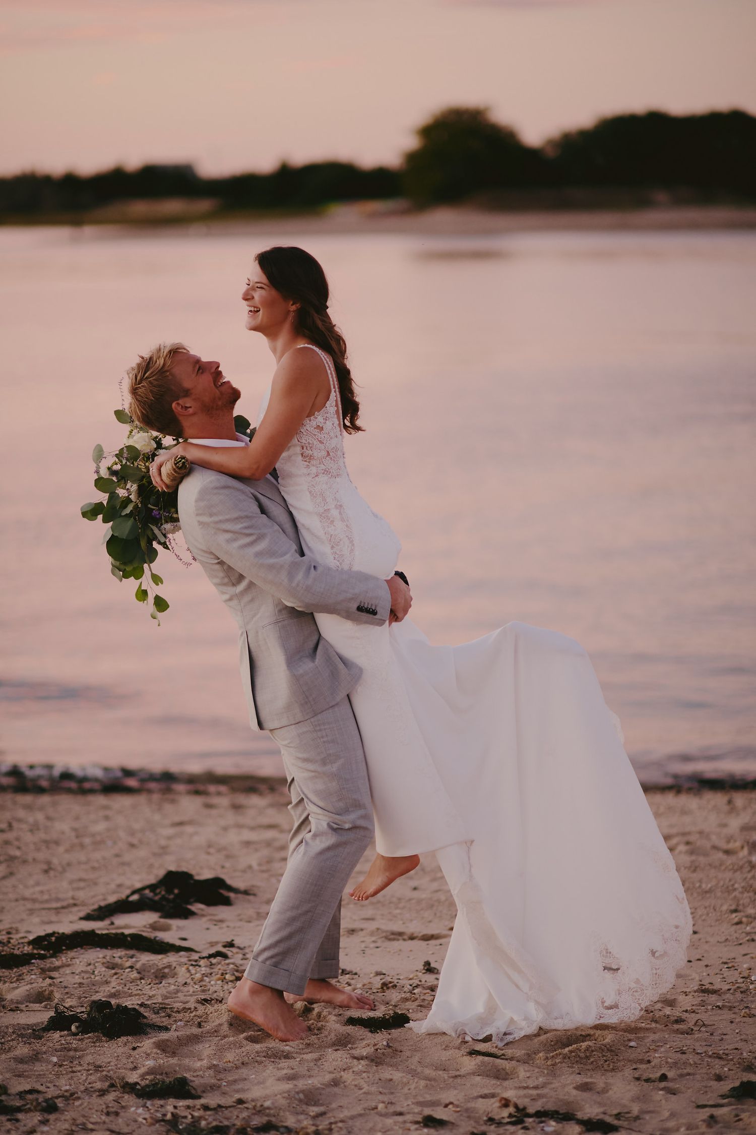 A couple in wedding attire share a romantic moment on a beach at sunset, with the bride holding a floral bouquet.