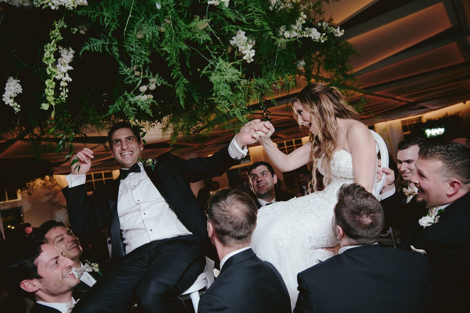 Wedding party celebrates with drinks at an indoor reception under lush green foliage and white floral decorations.
