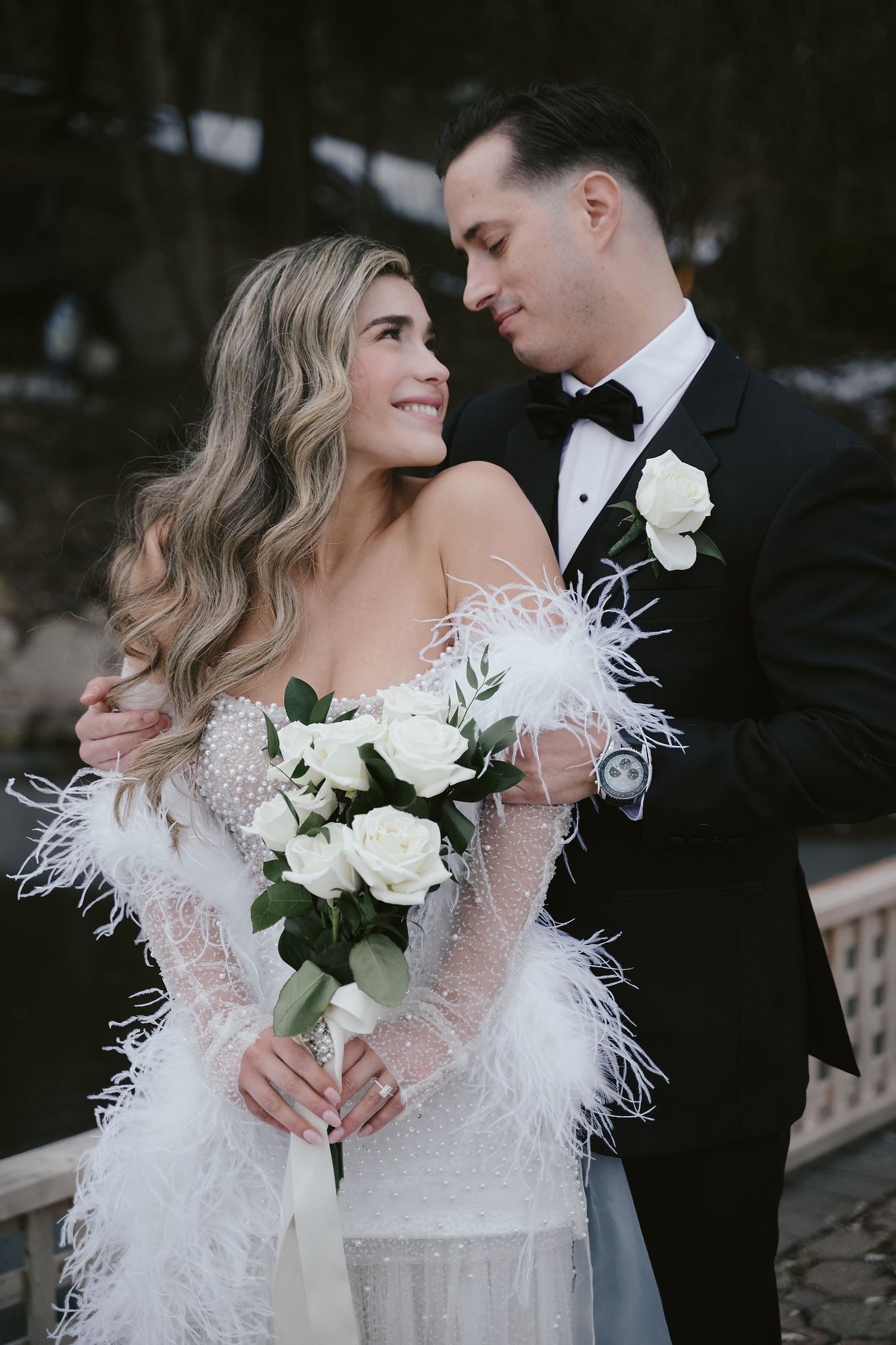 A couple in wedding attire share an intimate moment, with the bride holding a white rose bouquet with feather accents.