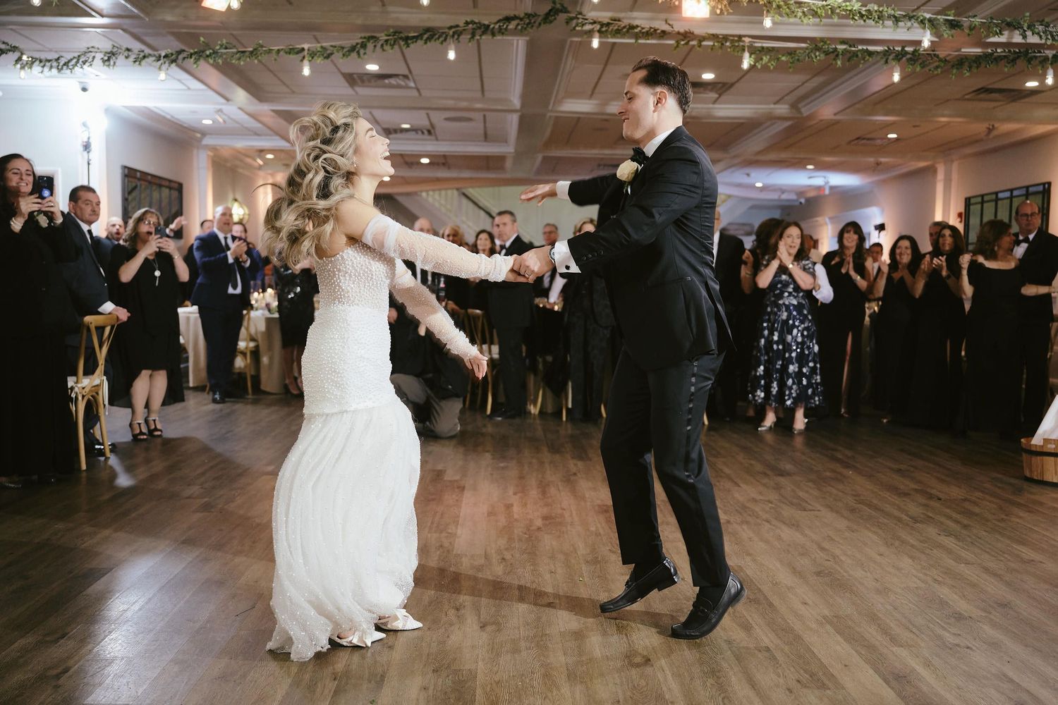 Bride and groom sharing their first dance on wooden floor surrounded by wedding guests.