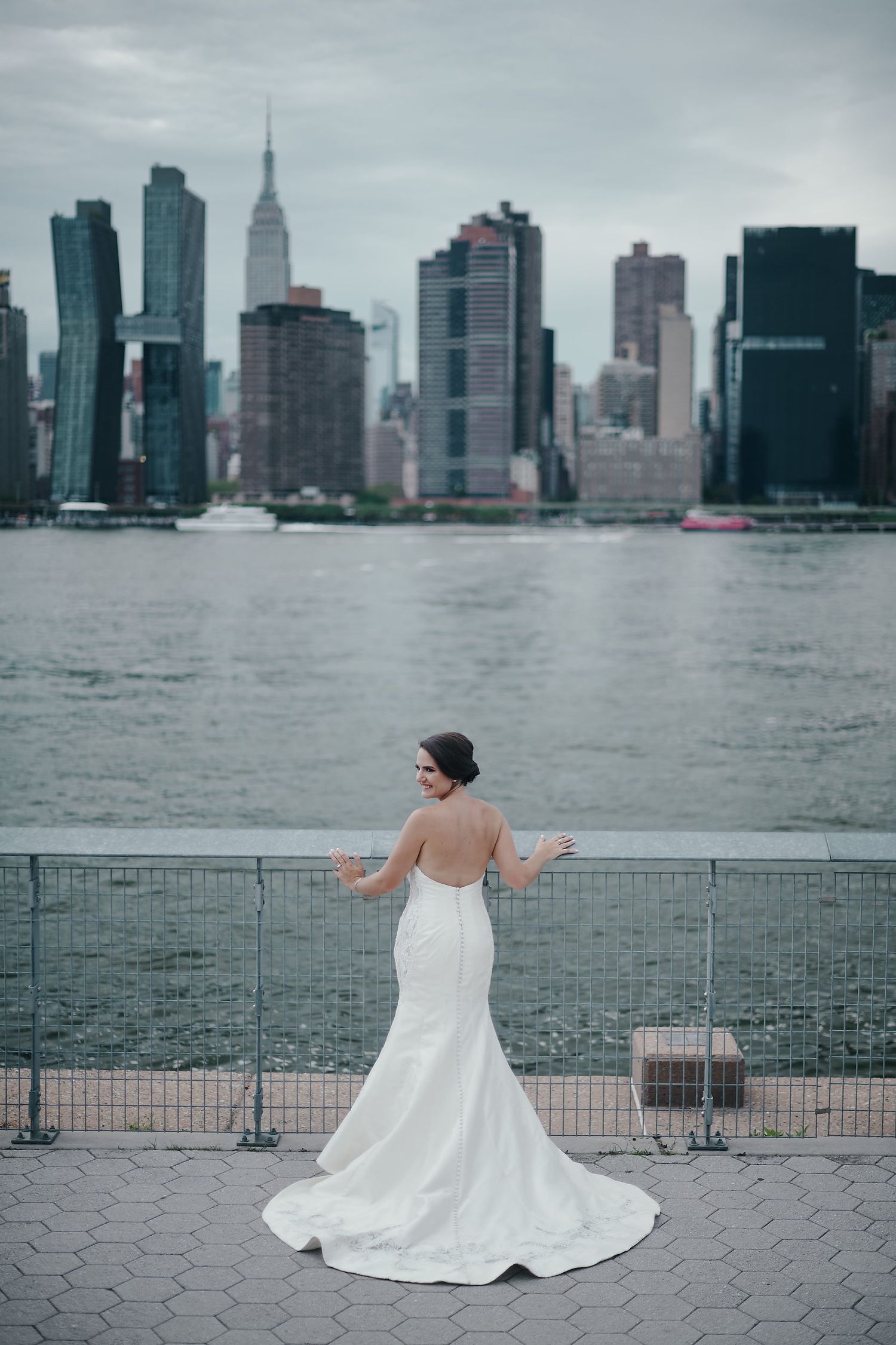 A bride in a white mermaid gown stands overlooking the New York City skyline on a cloudy day.
