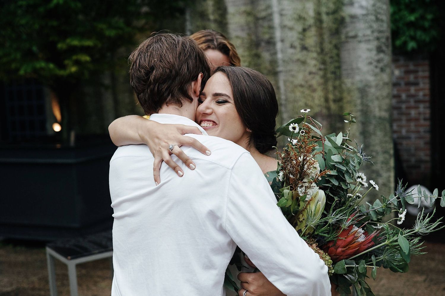 Two people embrace while holding a floral bouquet during an emotional outdoor wedding moment.
