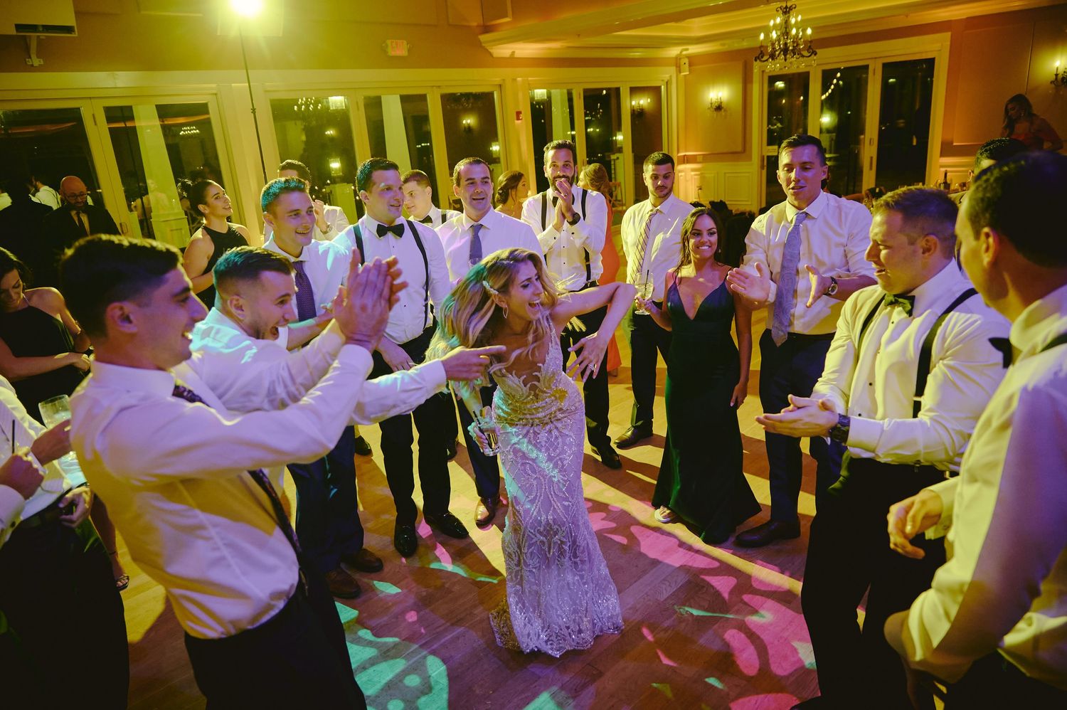 Wedding party forms a circle around the dancing couple at an indoor reception with warm yellow lighting.