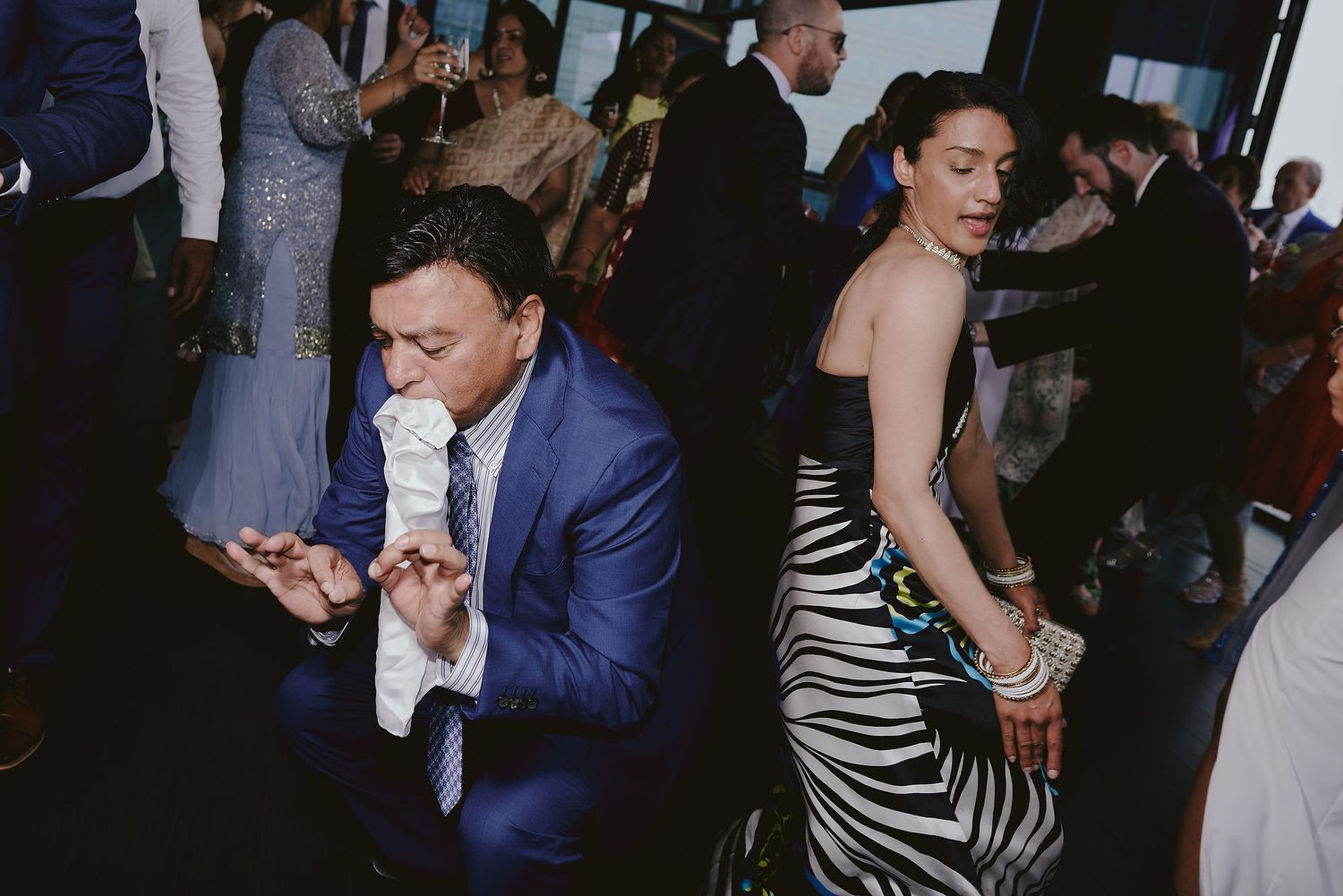 People dancing and singing at a lively wedding reception in a dimly lit venue.