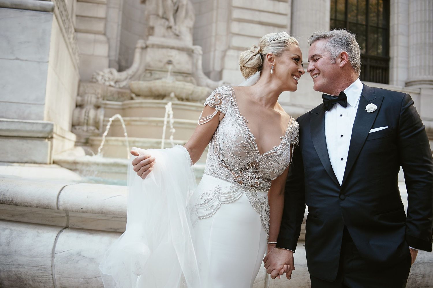 Elegant bride and groom share a tender moment outside historic stone building in formal wedding attire.