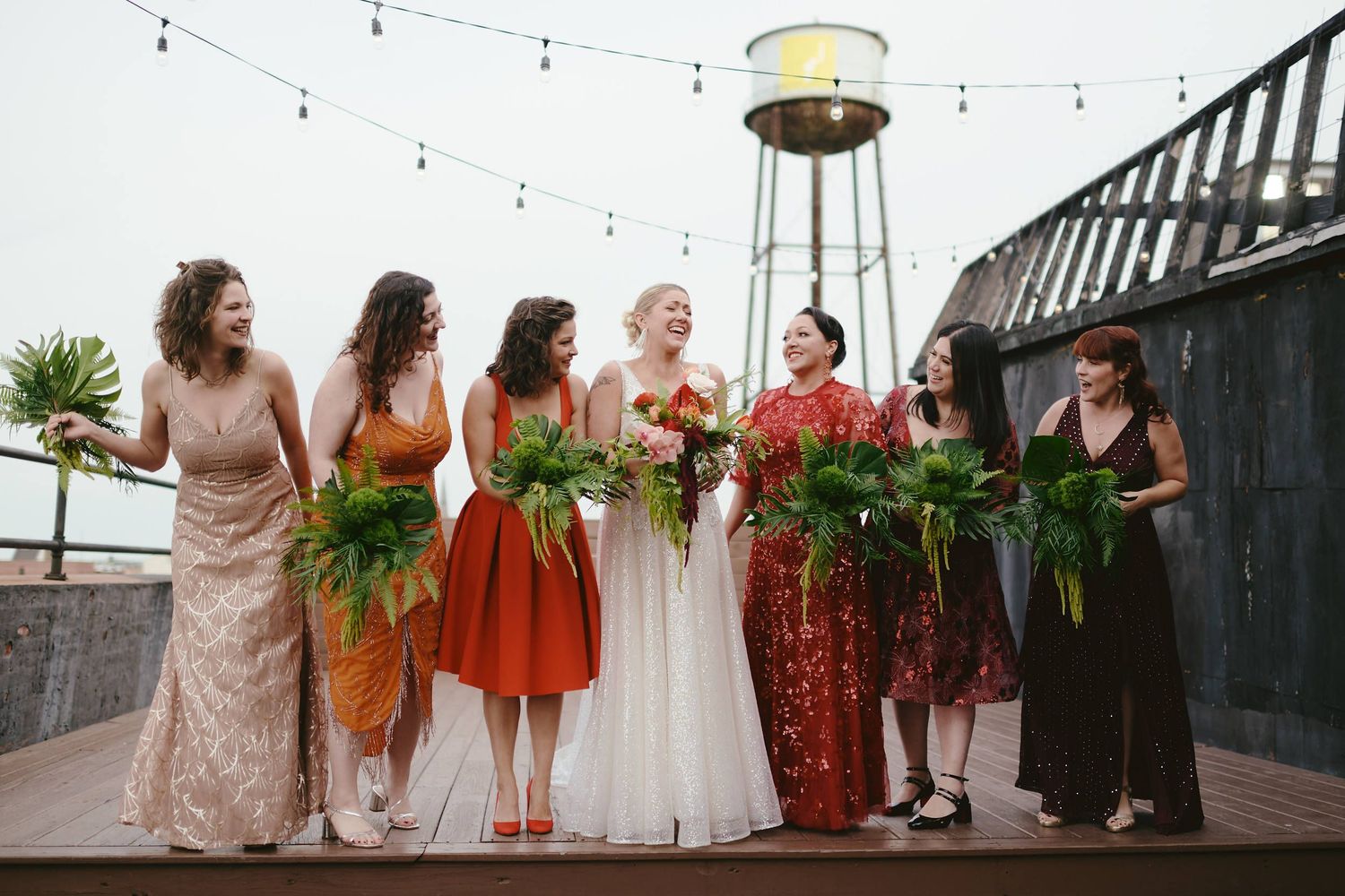 A bridal party in mismatched dresses stands on a rooftop with string lights and a water tower in the background.